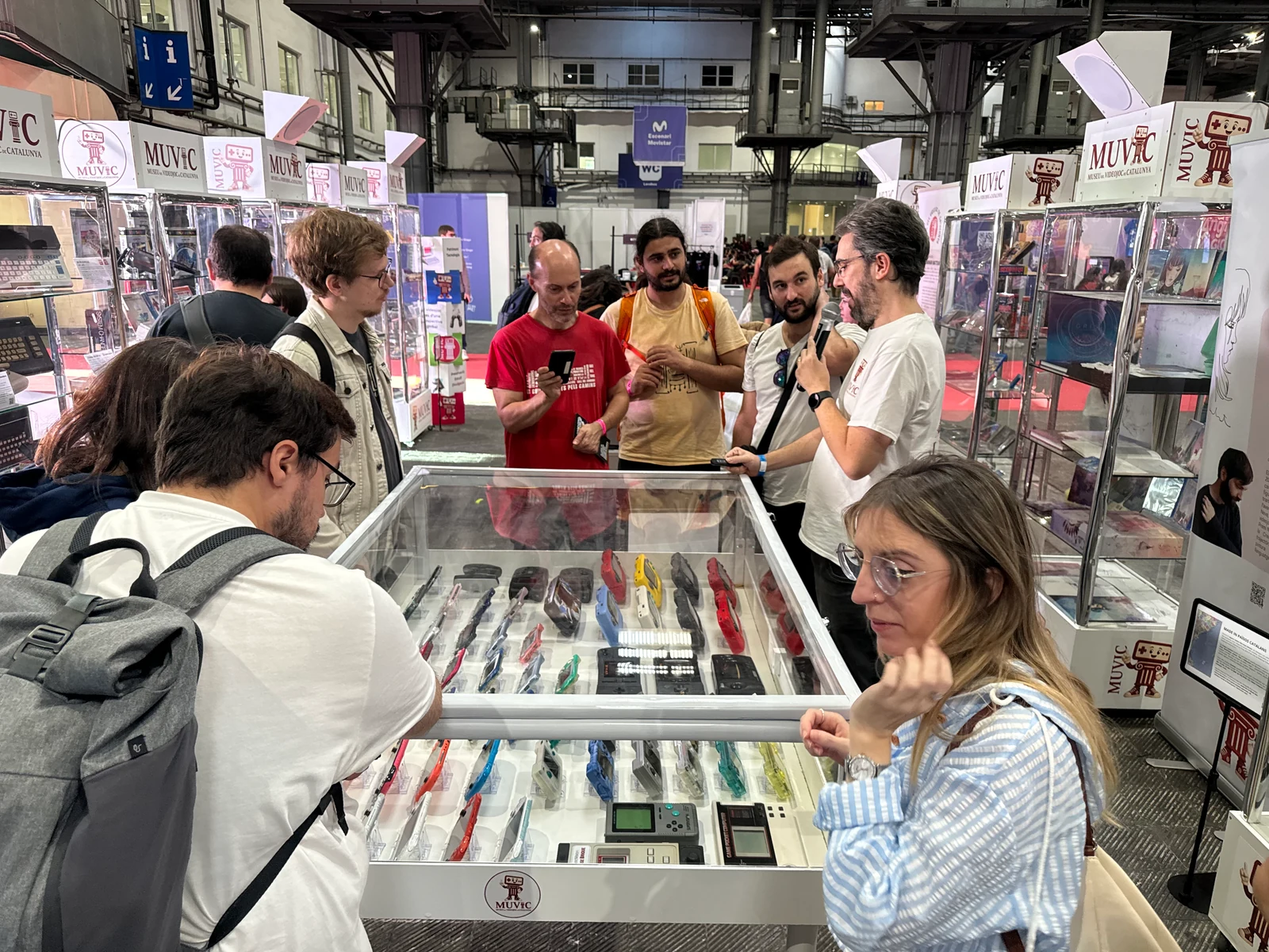 Visitants escolten una explicació de l’equip del MUVIC al costat d’una vitrina de consoles portàtils